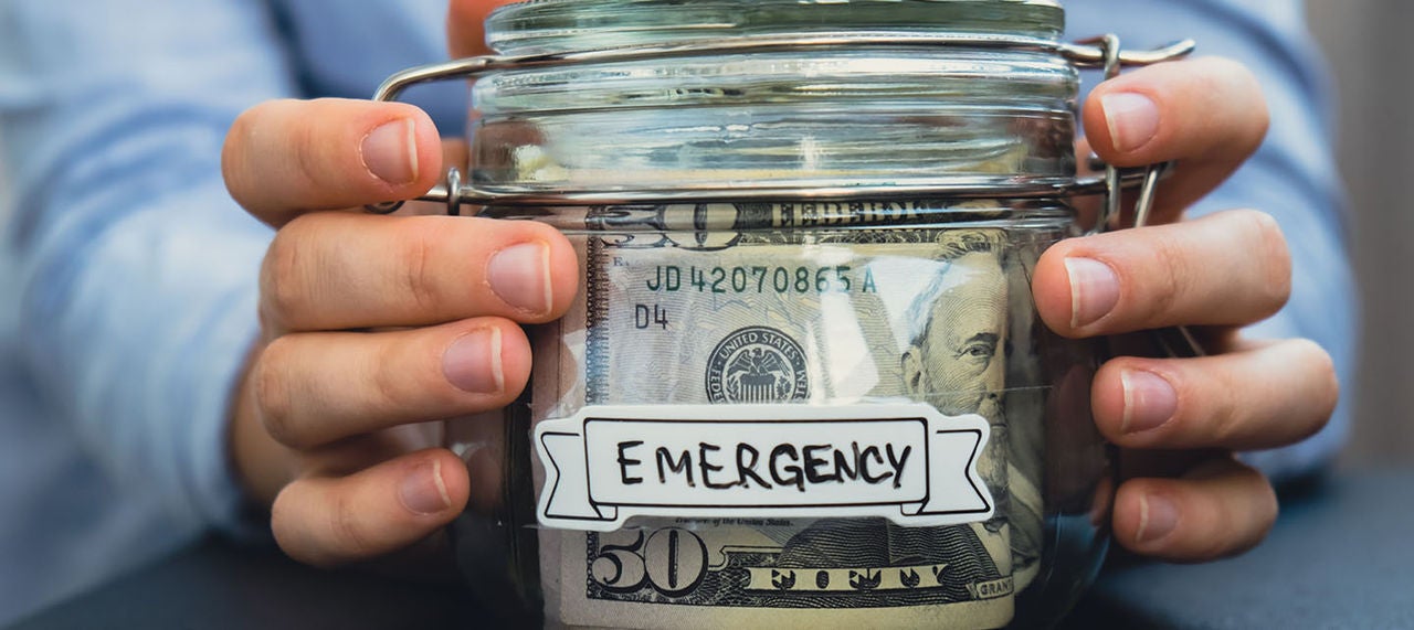 Hands holding a glass jar labeled 'Emergency' filled with dollar bills for savings