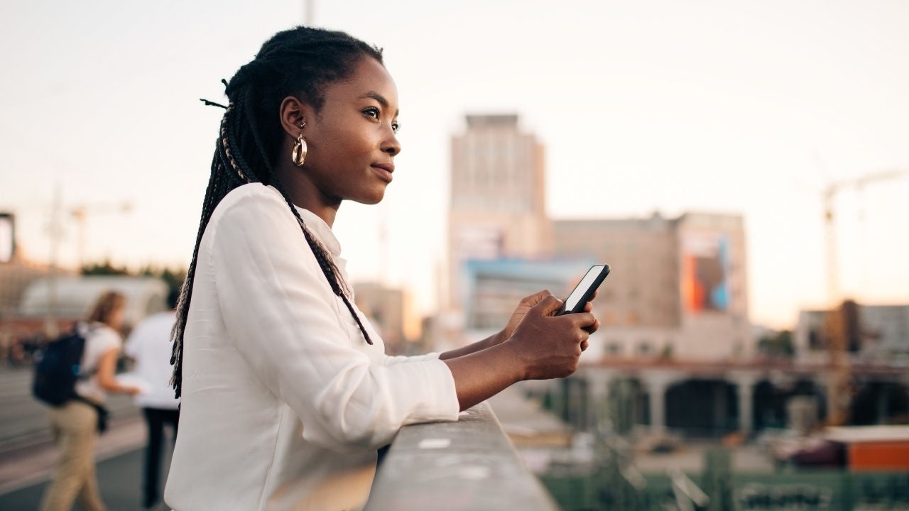 A woman holds her phone on a bridge looking into the distance in a city. 
