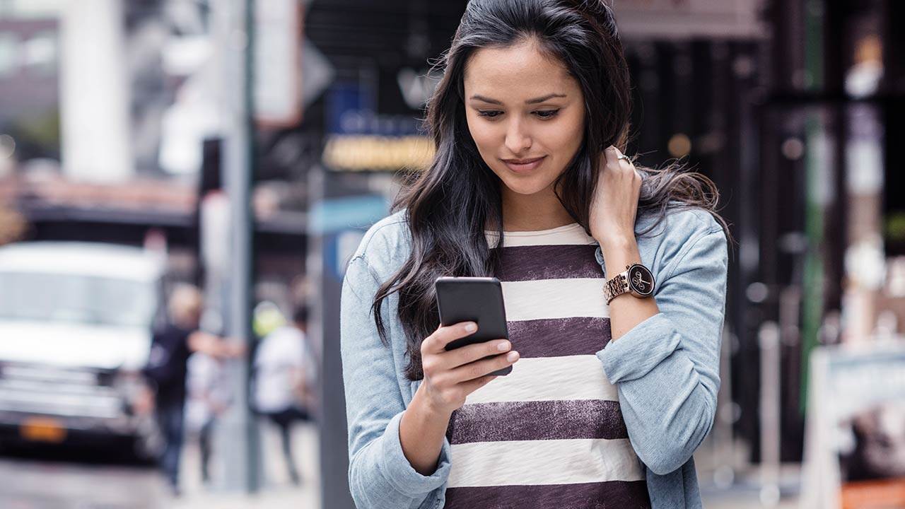Egy fiatal nő az utcán sétálva a mobiltelefonját használja.