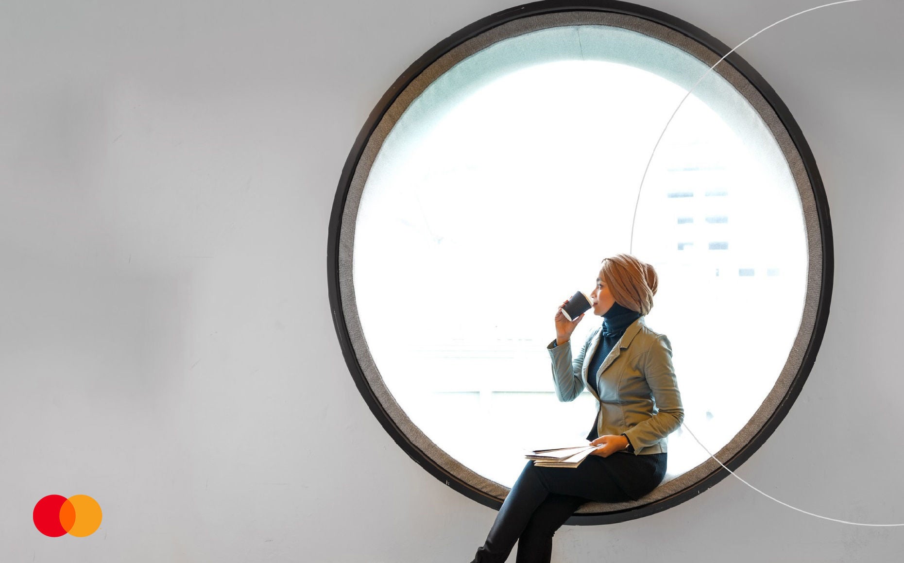 A woman in a light jacket sits in a large round window drinking from a takeaway cup, holding papers on her lap.