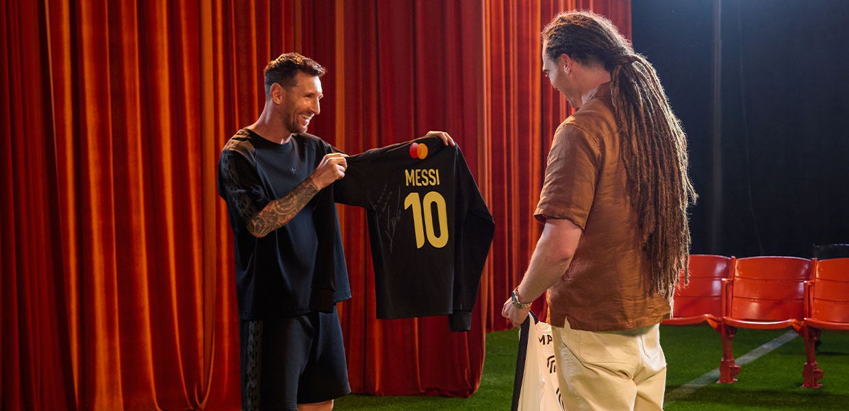 Lionel Messi smiling as he presents a signed black Messi #10 soccer jersey with a MasterCard logo to a male fan with dreadlocks during a promotional jersey exchange event.