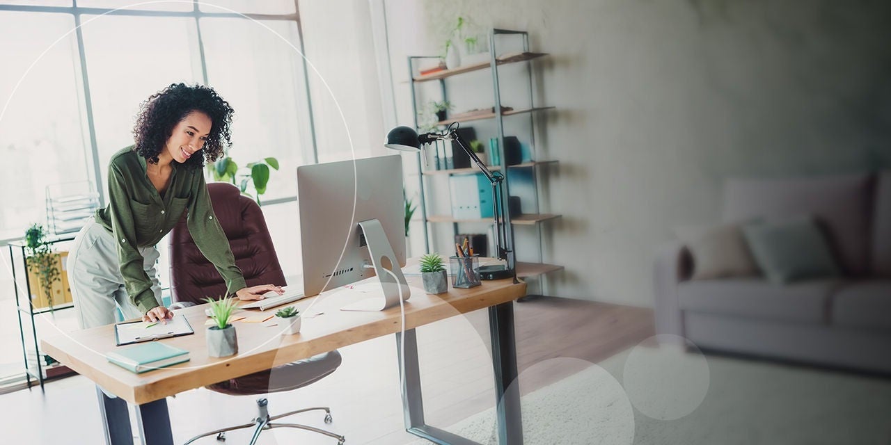 אישה מתכופפת מעל שולחנה כדי לבדוק פרטים מהמחשב שלה במשרד מואר, מקושט בעציצים קטנים