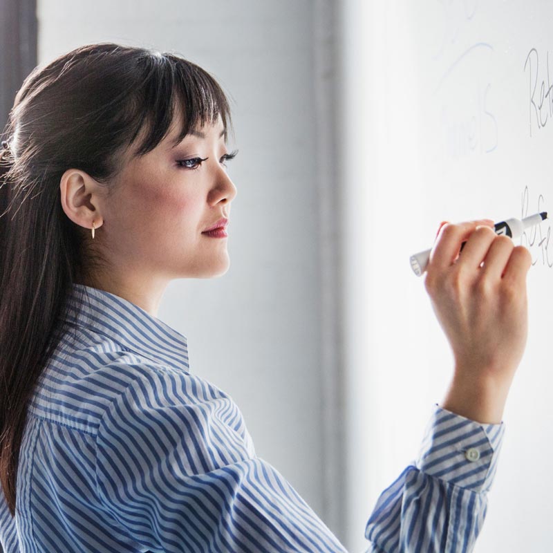 En person med mørkt hår og stribet skjorte skriver på en hvid tavle med en markør.