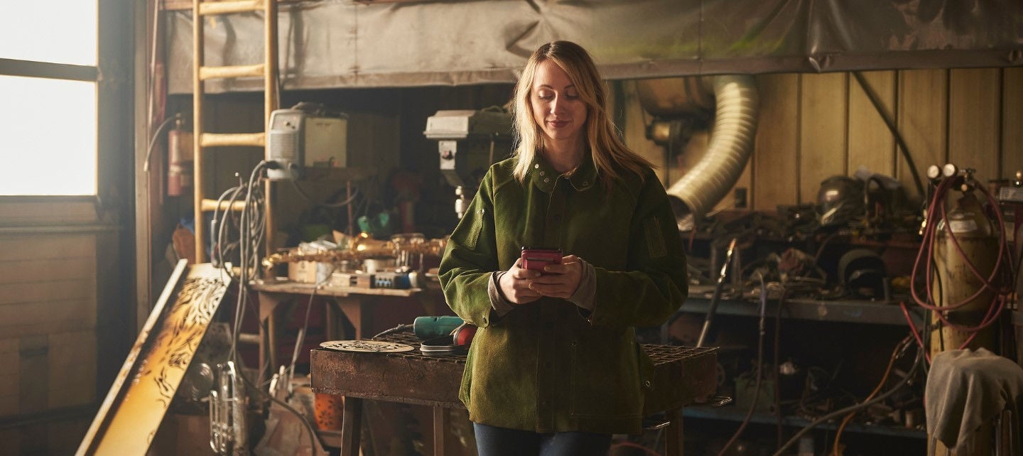A woman in a heavy work coat looks at her phone in a workshop. 