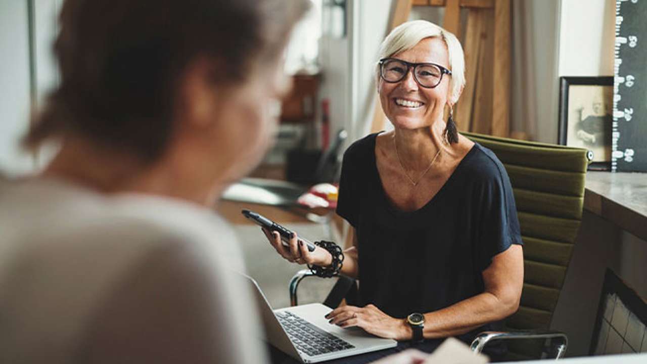 En ældre kvinde med blondt hår og briller sidder ved et bord med en computer og smiler.