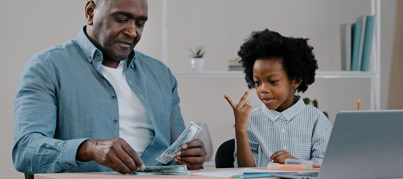 Father teaching young child about money while counting cash together at a table with a laptop nearby.