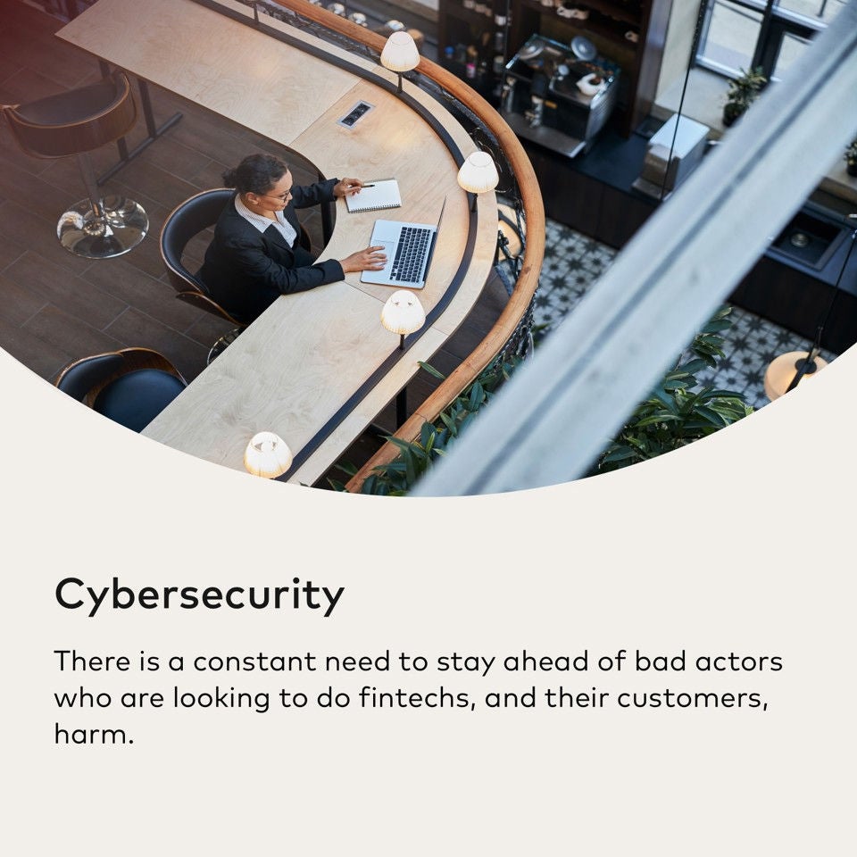 A woman in a suit sits at a curved wooden desk with a laptop and notebook.