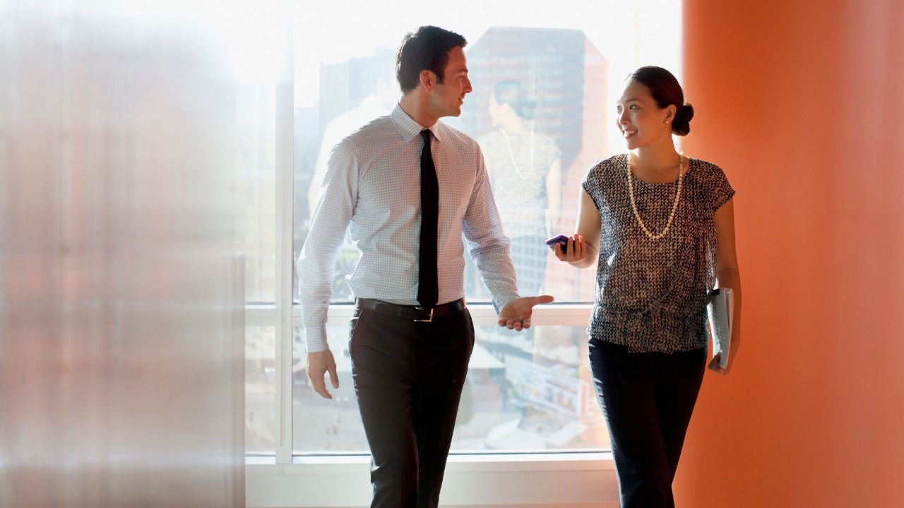 Two people walk down the hall in an office talking. 