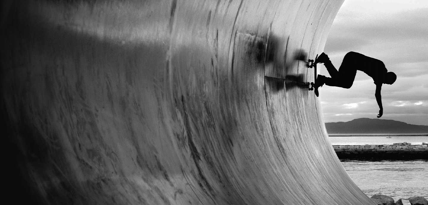 Et sort-hvidt billede af en skateboarder, der udfører et trick mod en buet væg, med hav og bjerge i baggrunden.