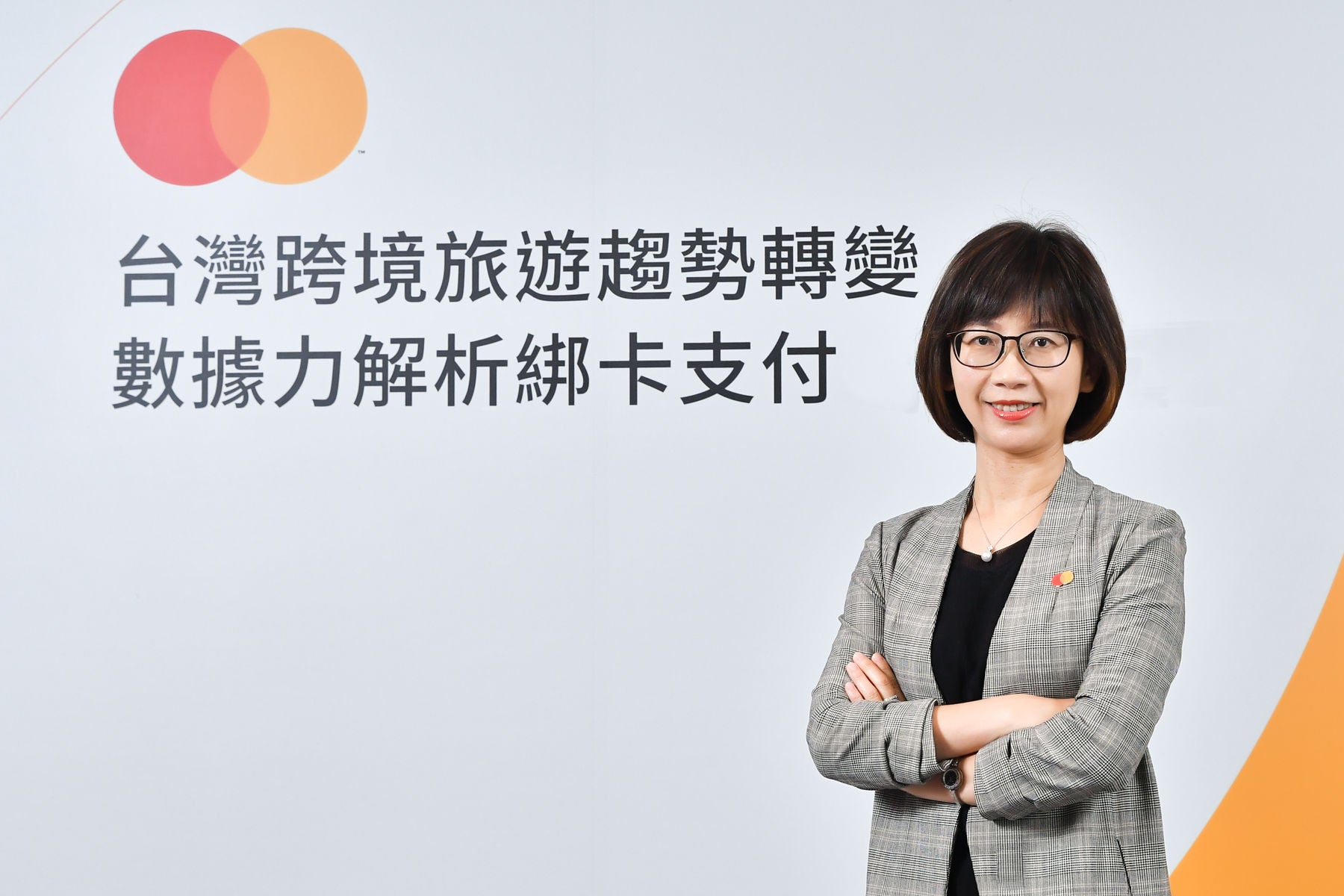 A woman in a grey checked blazer stands with her arms crossed, smiling in front of a white backdrop with Chinese text and a brand logo.