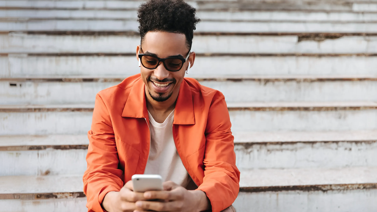 A man in an orange jacket and sunglasses on the steps looking at his phone