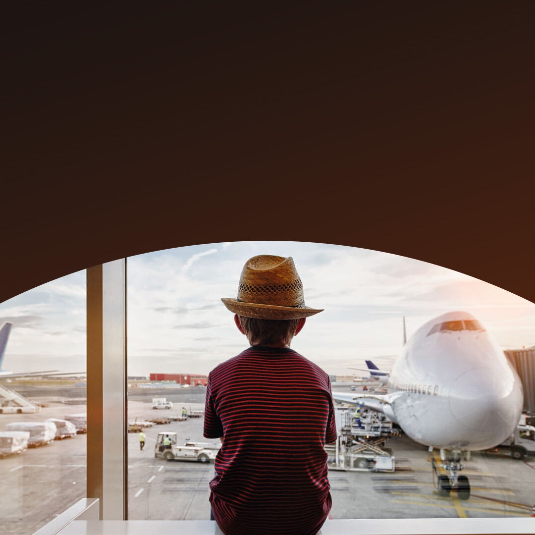A child wearing a hat sits by a large airport window, looking out at an airplane parked at the gate on the tarmac.