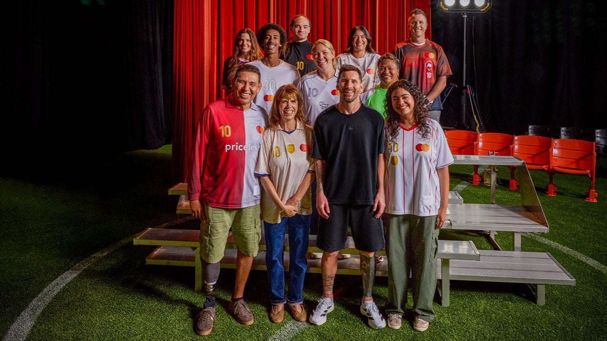 Lionel Messi posing with a diverse group of fans on a turf field stage set with red curtains. The group wears custom jerseys featuring MasterCard branding and number 10, highlighting a soccer fan event.