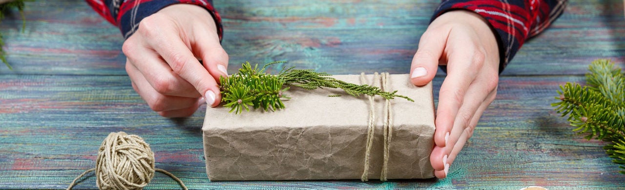 Kézzel csomagolt ajándék újrahasznosított papírba, természetes zsinórral átkötve és fenyőággal díszítve, rusztikus fa felületen.