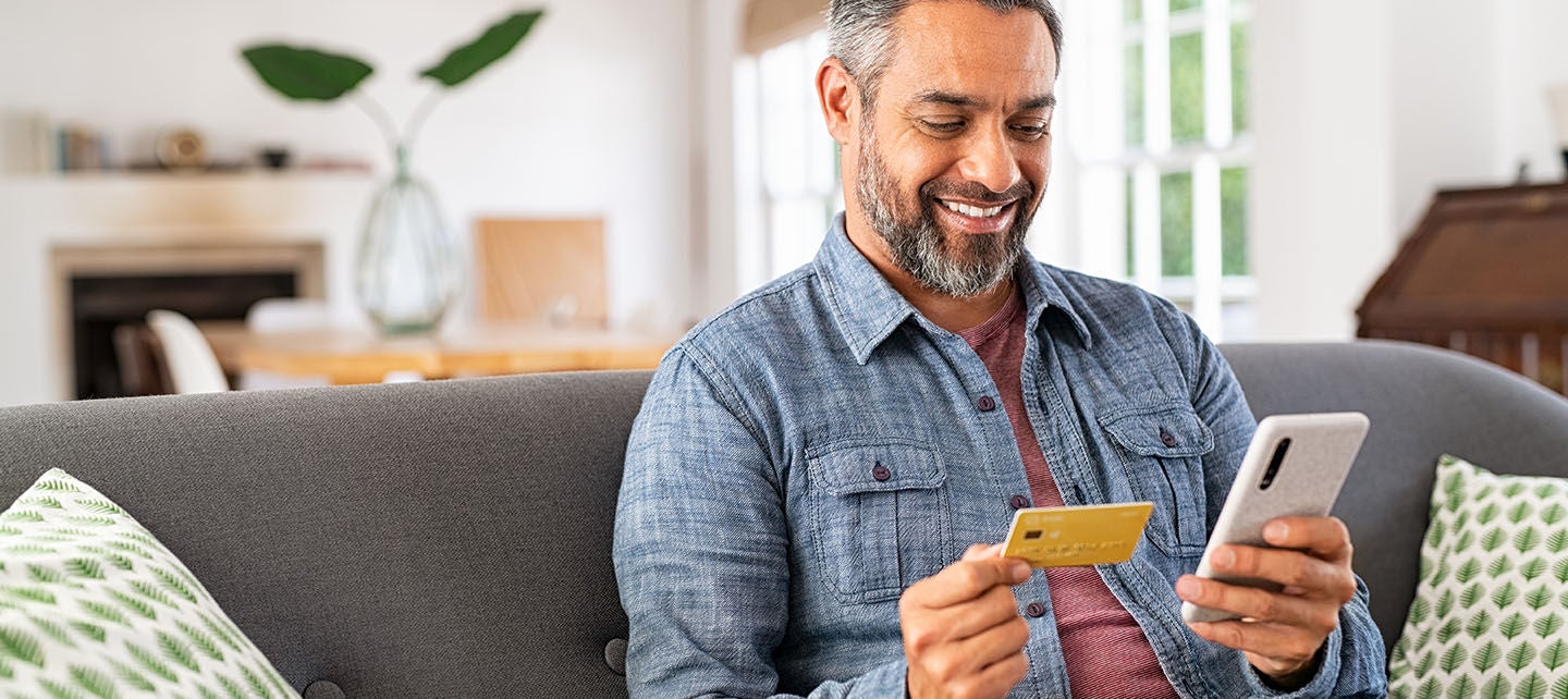 Middle-aged man sitting on a couch holding a credit card and smartphone for online shopping
