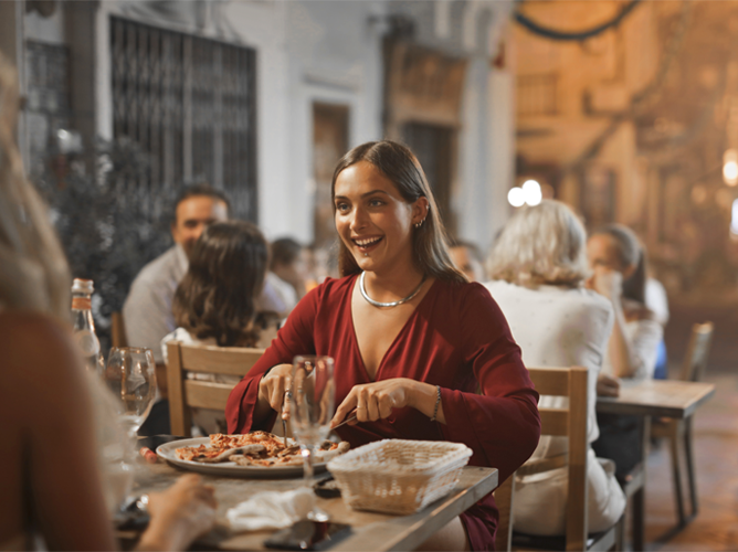 Smiling young woman in a red dress eating dinner at a lively outdoor restaurant, with friends in the background.