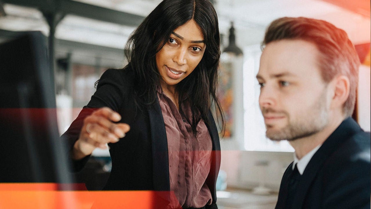 A woman points to a computer next to a man sitting at a desk. 