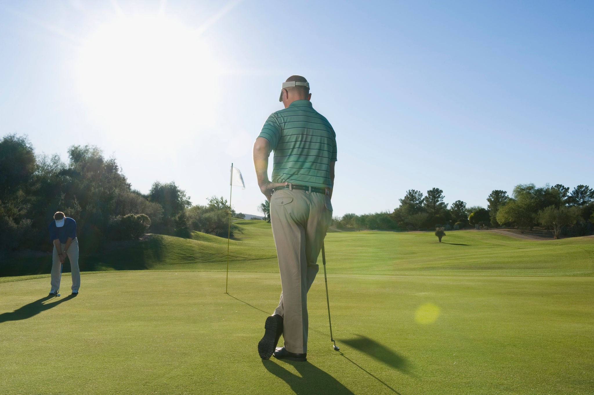 Two golfers on a sunlit green, one standing with a club and the other bending to putt, with a flag and trees in the background.