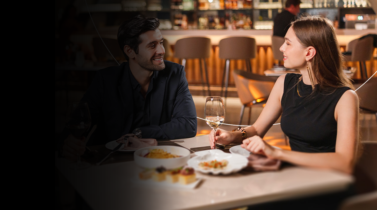 Dos personas conversan en una mesa de restaurante con copas de vino y comida servida.
