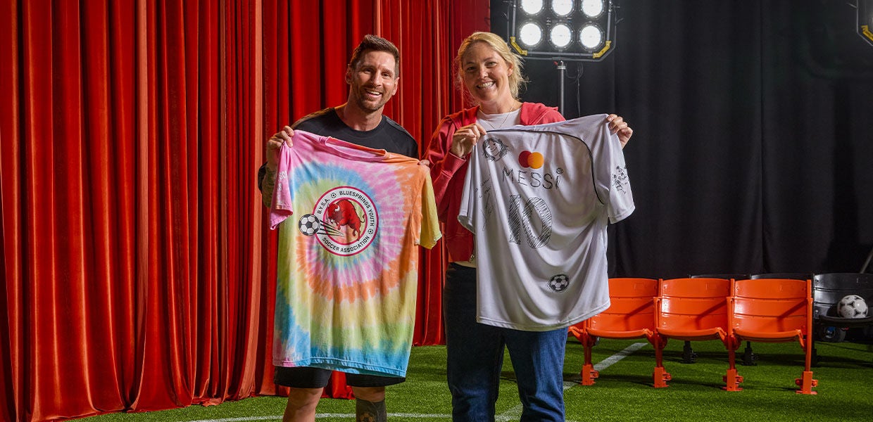Lionel Messi and a female soccer fan smiling while holding up exchanged jerseys. Messi holds a tie-dye Blue Springs Youth Soccer Association shirt, while the fan displays a white, autographed MasterCard Messi #10 jersey against a red curtain background.