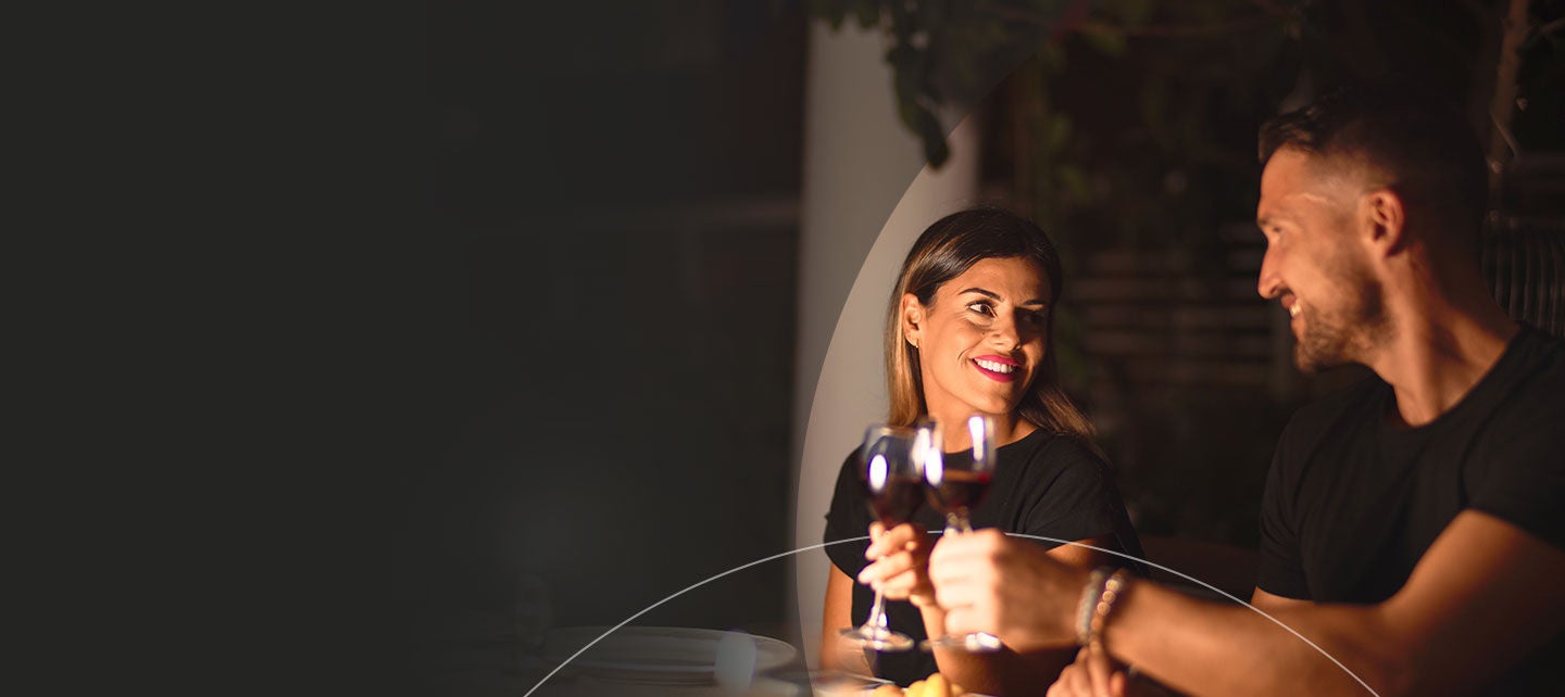 Man and woman sitting at a table looking at each other clinking their glasses of red wine.