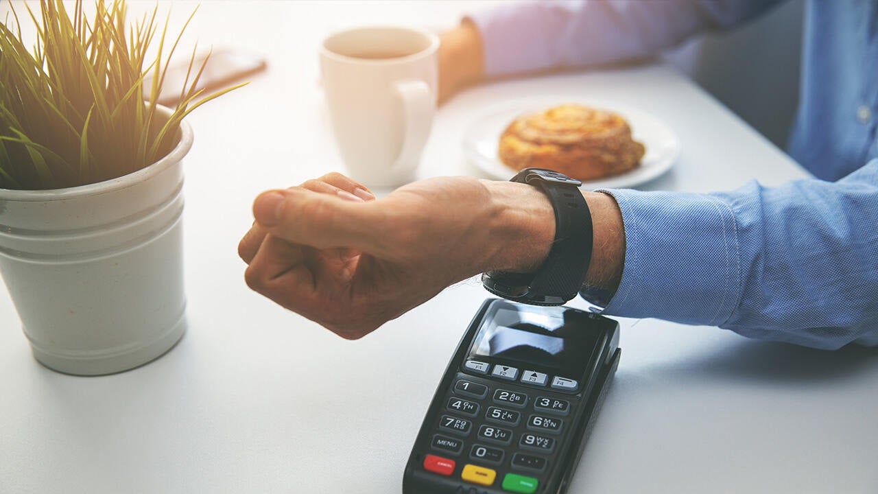 Okosórás érintéses fizetés kávézóban bankkártya-terminálnál, asztalon kávé és sütemény mellett.
