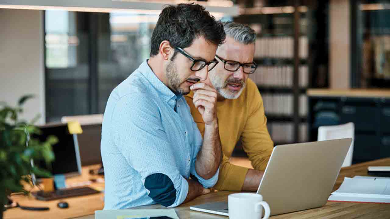 Két férfi egy modern, letisztult irodai környezetben közösen dolgozik egy nyitott laptop előtt. Az előtérben egy fiatalabb férfi látható világoskék ingben, szemüveggel, enyhén előrehajolva, gondolkodó testtartásban.