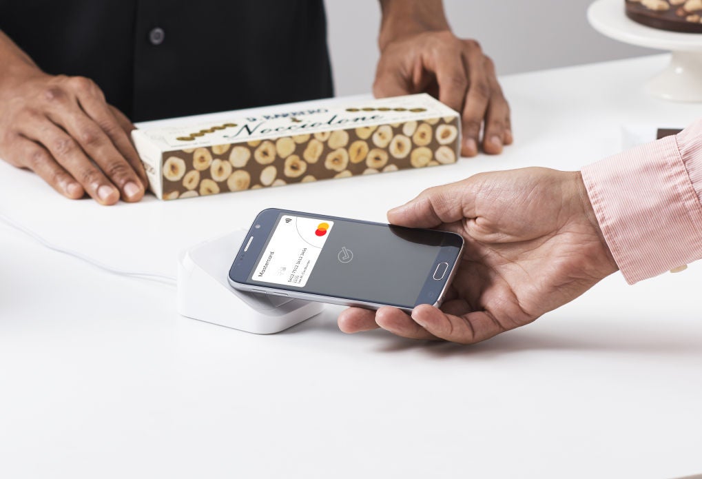 A close-up of a hand holding a phone up to a small white contactless terminal on a counter, with a boxed product and a shop assistant behind.