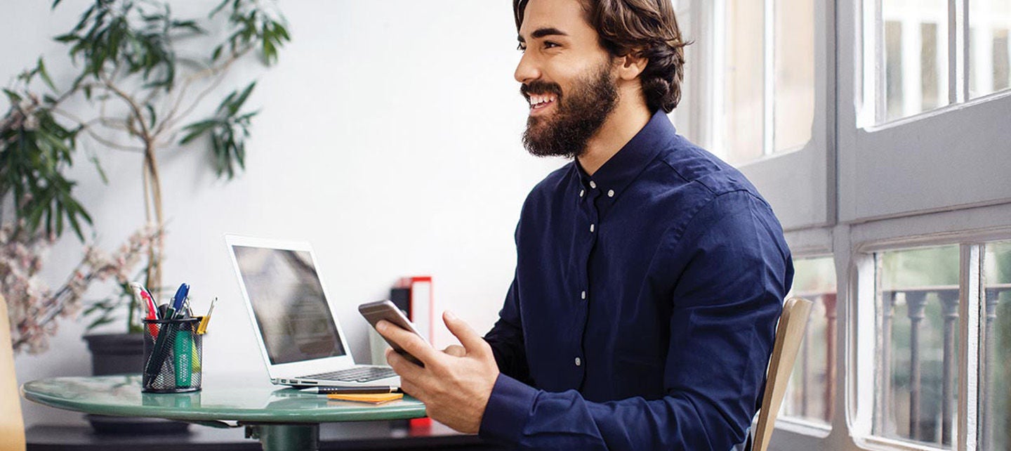 En person som smiler mens de holder en mobiltelefon og sitter ved et bord med en bærbar datamaskin.