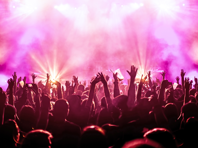 A large, excited crowd at a concert with hands raised toward the stage lights, illustrating the economic boost from live events.
