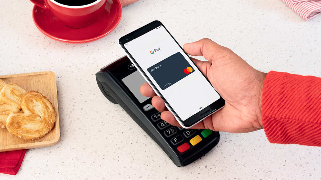 Okostelefonos érintéses fizetés Google Pay-jel kávézóban bankkártya-terminálnál, kávé és péksütemény mellett.