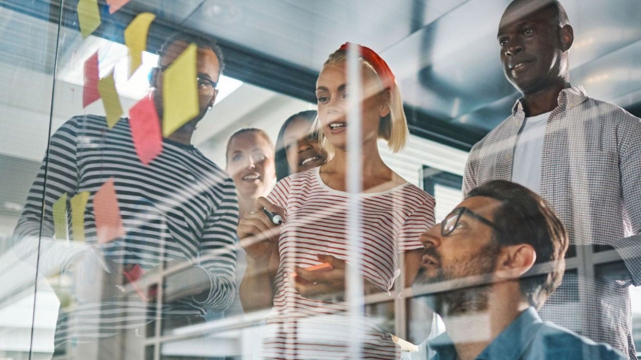 A group of employees look at Post-Its on a clear wall in an office. 
