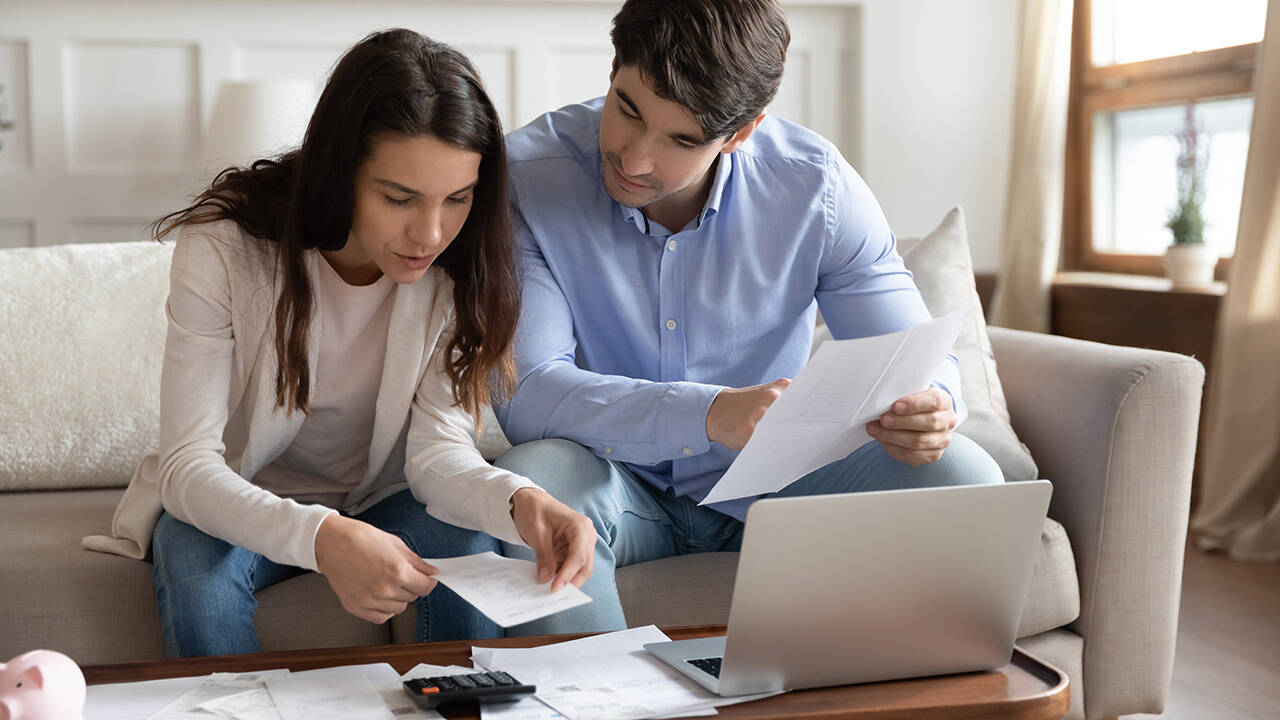 Fiatal pár a kanapén ülve számlákat és papírokat néz át laptop és számológép mellett, miközben közösen intézik a pénzügyi teendőket.