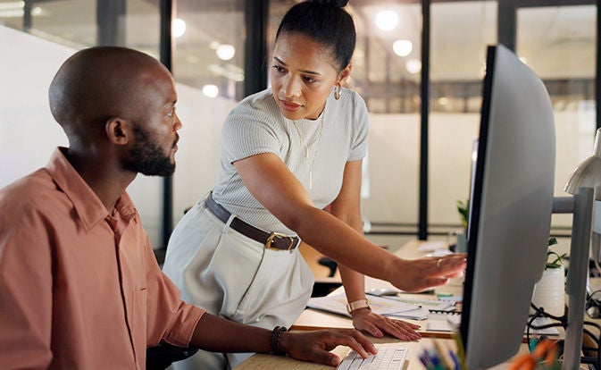 Two colleagues collaborate at a desk, reviewing content on a computer screen in a modern office setting.