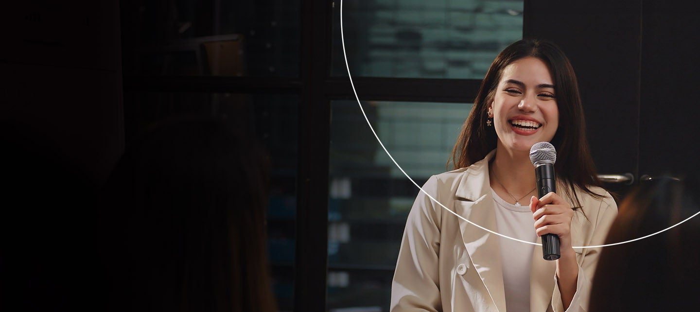 Čovjek stoji i gestikulira dok prezentira trojici ljudi koji sjede u modernom uredu sa staklenim zidovima i policama u pozadini.