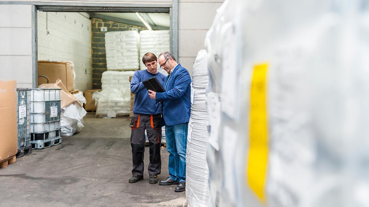 Két raktári dolgozó egy ipari raktárban táblagépen ellenőrzi az árukészletet a raklapokra csomagolt termékek között