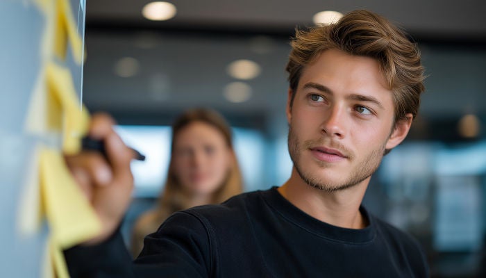 A man with light hair writes on yellow sticky notes on a wall in an office setting. A woman is blurred in the background. The atmosphere is focused.