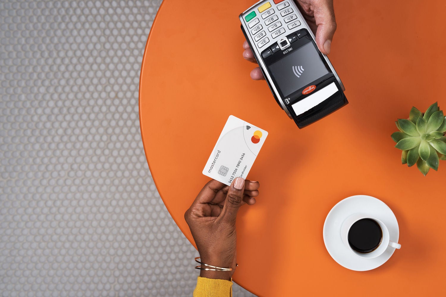 An overhead view of a hand holding a white payment card up to a card terminal held by another hand, on an orange table with a coffee cup and a succulent.