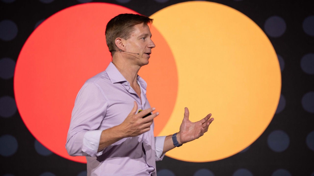 Greg Ulrich on stage at RiskX in front of the Mastercard logo. 