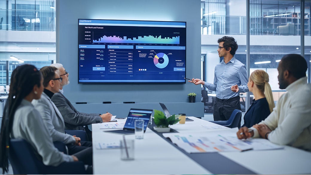 Modern irodai prezentáció, ahol egy férfi egy nagy képernyőn látható színes adatgrafikonokat és dashboardot elemez a munkatársai előtt.