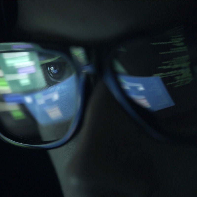 A man in a dark room with computer coding reflected in his eyeglasses. 