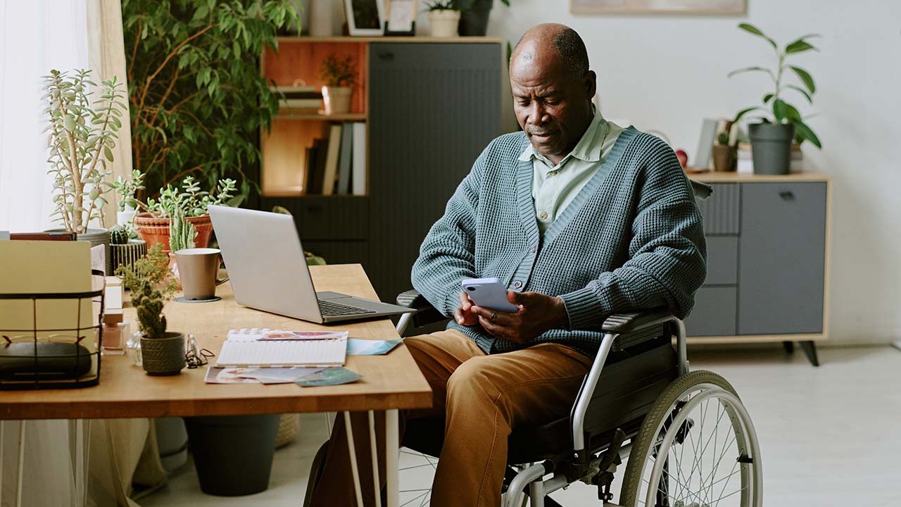 Man in wheelchair using phone and laptop in home office.
