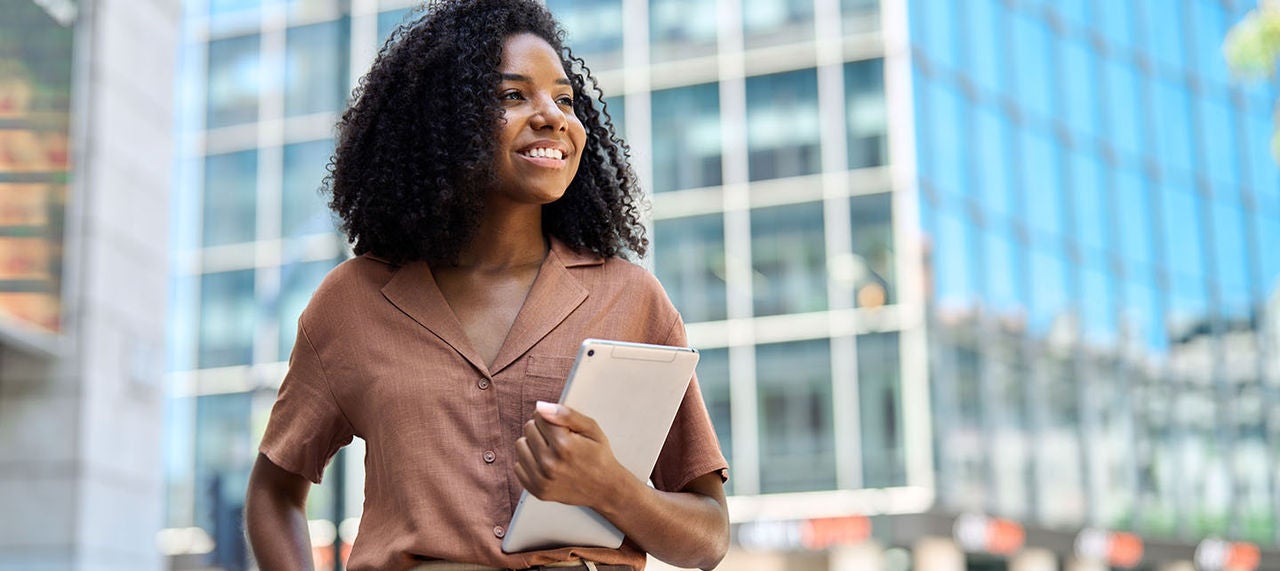 Confident young woman holding tablet and smiling in front of modern office buildings.