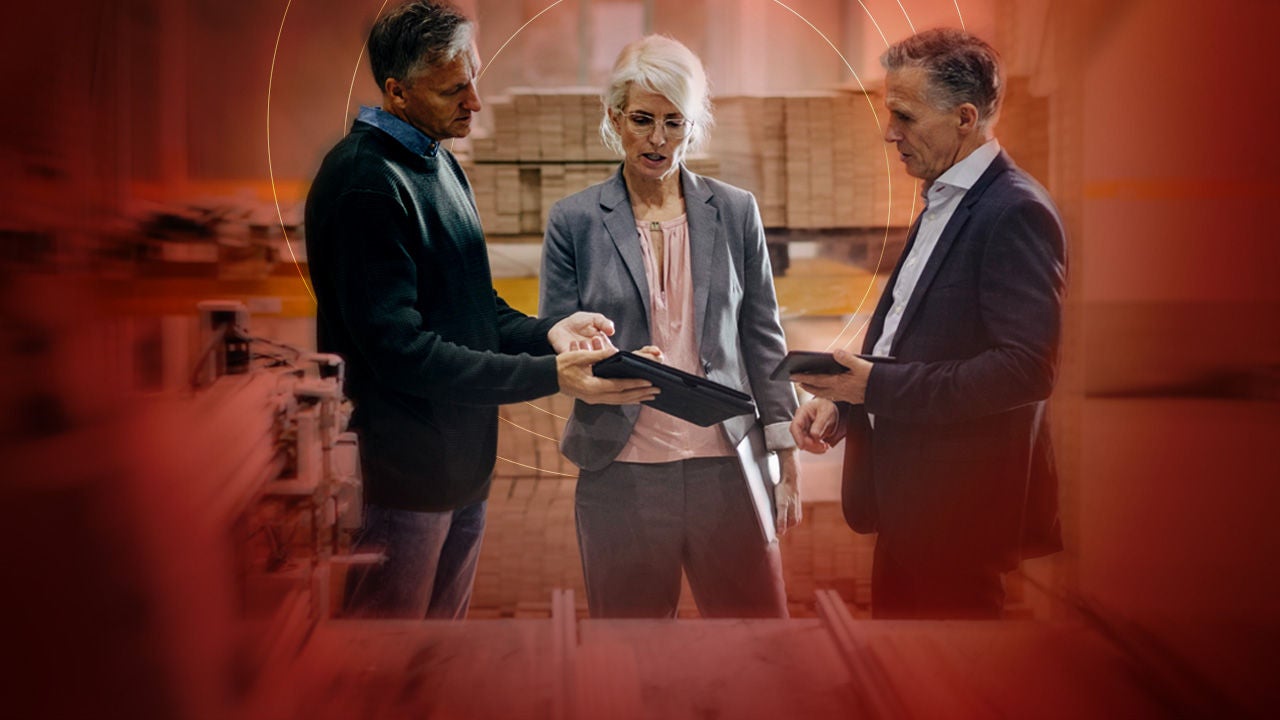 Three business professionals, two men and one woman, collaborating in a warehouse setting, reviewing information on tablets.