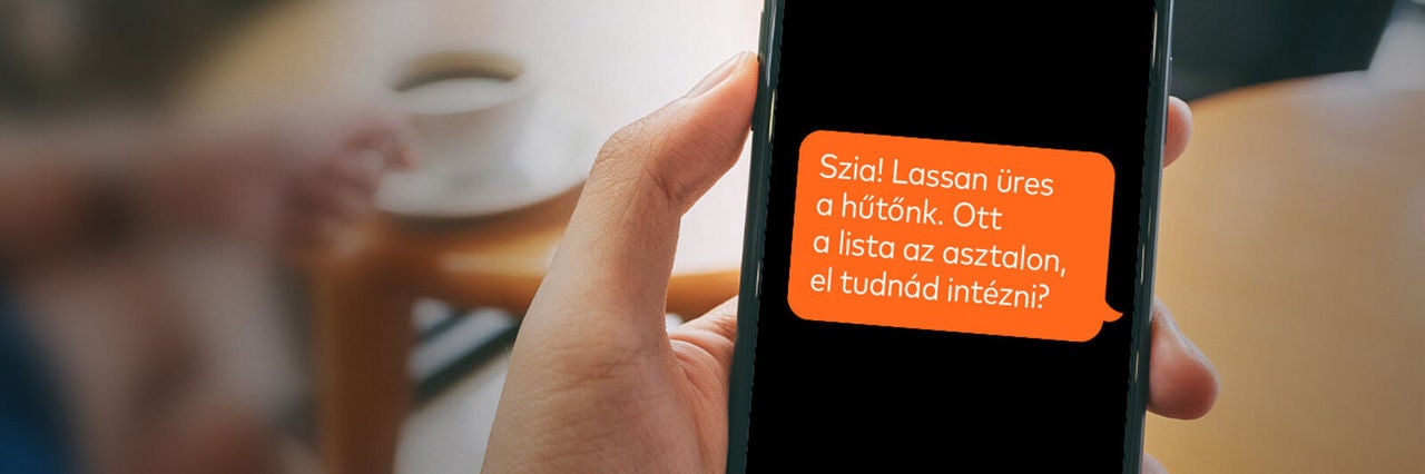 Okostelefon kijelzőjén üzenet a bevásárlás elintézéséről, kézben tartva, elmosódott otthoni háttérrel.