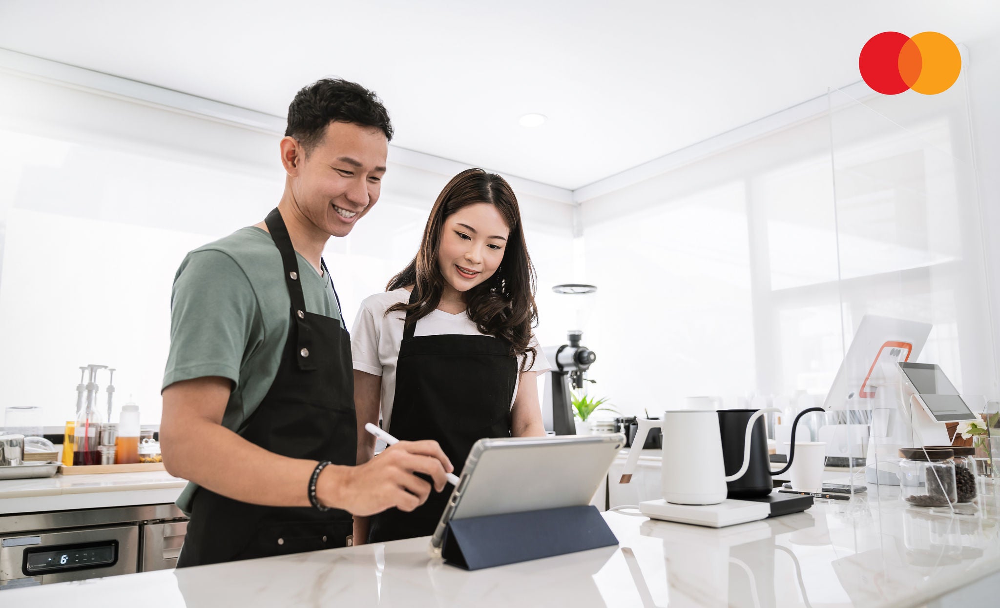 A man and a woman in black aprons stand behind a café counter looking at a tablet together, with coffee equipment and a payment terminal around them.