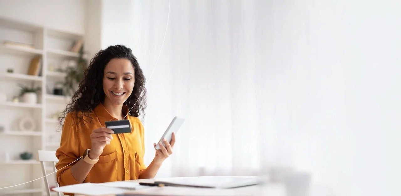 אישה יושבת ליד שולחן, אוחזת בטלפון שלה ומסתכלת על כרטיס האשראי שלה.