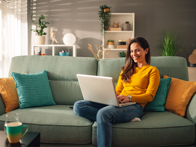A smiling woman works comfortably on her laptop from a sofa in her living room, representing the modern blend of clicks and commutes.