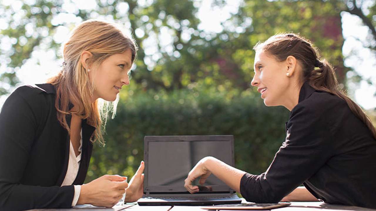 To kvinder sidder ved et bord udendørs og arbejder på en bærbar computer.