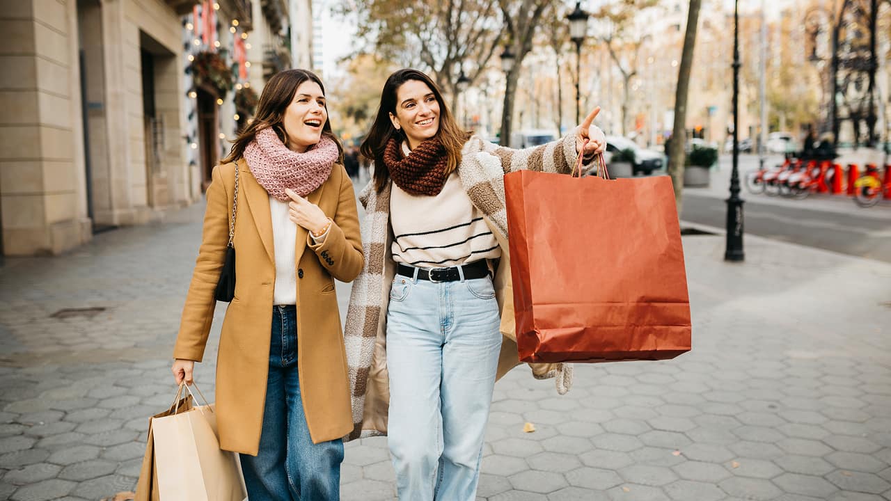 două femei mergând pe o stradă a unui oraș ținând în mână sacoșe de cumpărături
